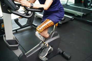Young female with one prosthetic leg with exercising with a spinning bike in the gym to practice...