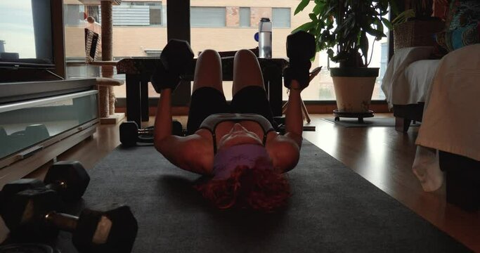 A redhead woman lying on the floor training her chest with dumbbells.