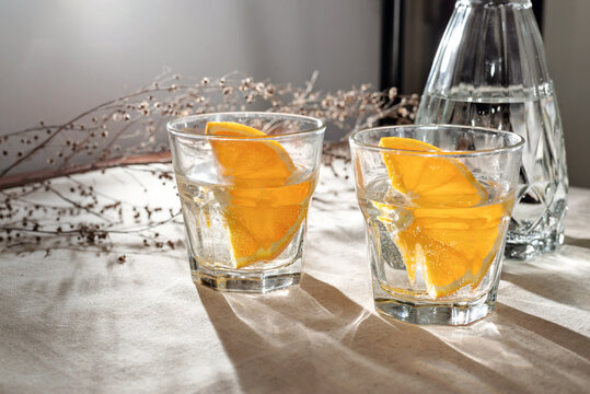 Glasses With Orange Infused Water And Bottle On Beige Table With Aesthetic Sunlight Shadows, Lifestyle Healthy Drink, Alcohol Cocktails Bar Still Life