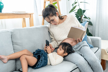 In a cozy living room, A big belly pregnant Asian mother and her little daughter spend their free time together happily. A mother look interesting at daughter's smartphone while she's reading book.