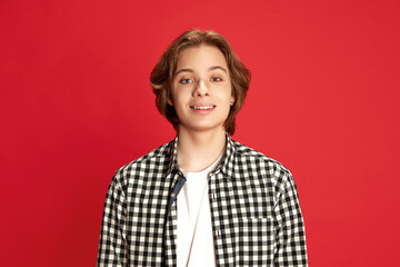 Young teen boy with long hair wearing checkered shirt looking at camera with smile over red studio...