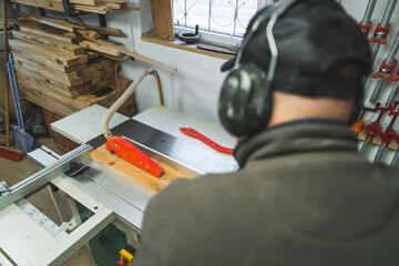 Back view of professional carpenter in protective earphones using table saw blade to part a piece of wood. Blurred silhouette in the foreground. High quality photo