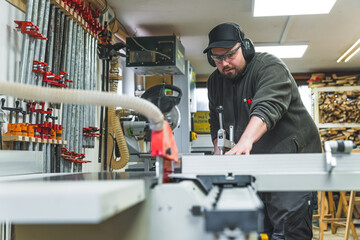 Obraz na płótnie Canvas Focused professional male craftsman saws wood on a circular saw in protective eyewear and headphones. Woodworking studio interior. High quality photo