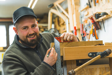 Smiling positive male carpenter using measuring bracket and pencil to create another wooden art...