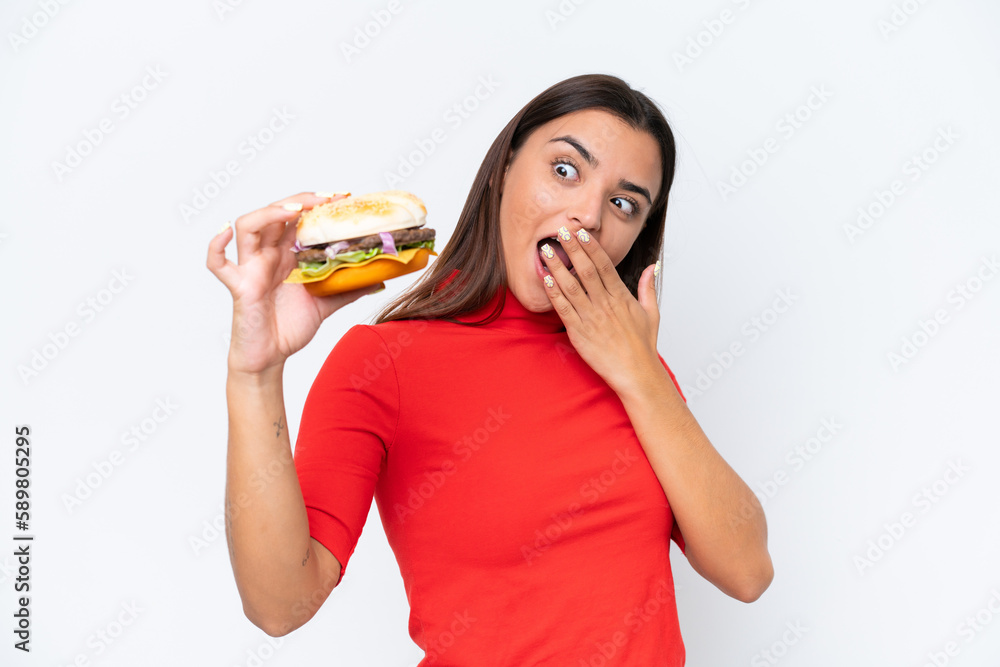 Wall mural Young caucasian woman holding a burger isolated on white background with surprise and shocked facial expression