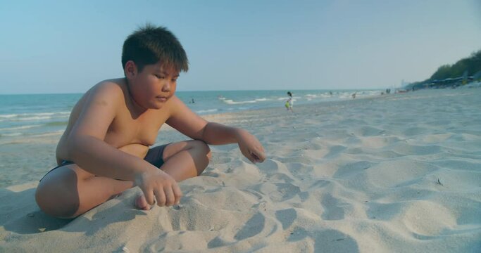 (Slow motion) A happy Asian boy who had a rather chubby figure, enjoying and playing in the sand on the sea beach in the evening sunset time.