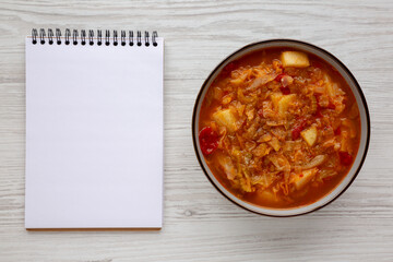 Homemade Shredded Cabbage and Potato Soup in a Bowl, blank notepad, top view. Overhead, from above, flat lay. Copy space.