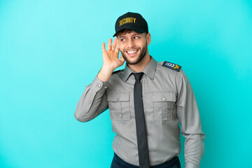 Young security man isolated on blue background listening to something by putting hand on the ear
