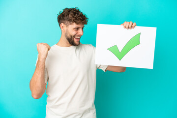 Young handsome caucasian man isolated on blue background holding a placard with text Green check mark icon and celebration a victory