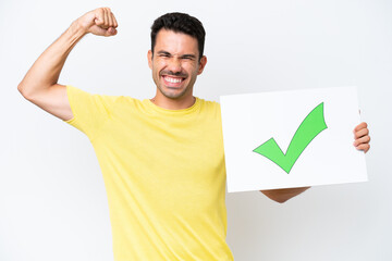 Young handsome man over isolated white background holding a placard with text Green check mark icon and doing strong gesture