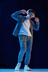 Full-length image of young guy in casual clothes listening to music in headphones against dark background in neon light. Enjoying sounds. Concept of human emotions, youth, fashion, lifestyle