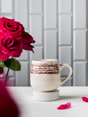 Fresh tea in a light-colored ceramic mug on a white wooden table. Roses in the background. Rose petals on the table. gentle morning. Tea drinking. Copy space