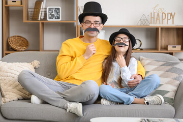 Portrait of father and his little daughter with paper mustache sitting on sofa at home