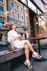 Smiling woman drinking coffee on the veranda of a cozy cafe. Tourist woman enjoys architecture and irrigating weather of city streets with a cup of coffee. Tourism, travel and vacation concept.