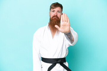 Redhead man with long beard doing karate isolated on blue background making stop gesture