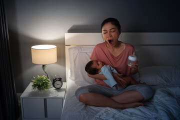 sleepy mother yawning and trying to feeding milk bottle to her crying newborn baby on bed at night
