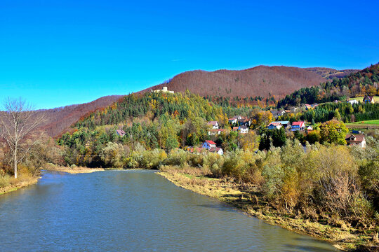 Autumn mountains landscape. Trees on a slope with dry grass and wooded mountains under blue sky and Poprad river near Rytro Village, Beskid Sadecki, Poland.