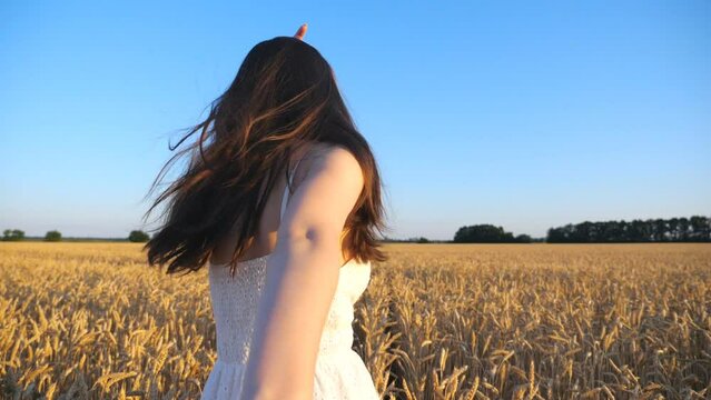 Woman in white dress holding male hand and running through large wheat field. Happy girl leading her boyfriend among yellow crop ears looking back into camera. Freedom or summer concept. POV Slow mo
