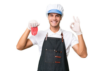 Butcher caucasian man wearing an apron and serving fresh cut meat over isolated background showing ok sign with fingers