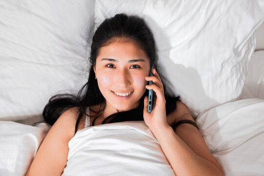 Top view of adorable happy cheerful smiled asian korean japanese kazachstan girl laying in bed waking up in the morning having call from someone talking on phone wth friends looking at camera.
