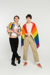 full length of young nonbinary models in stylish clothes posing with rainbow flags on grey background.