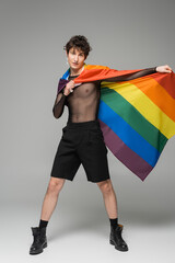 full length of brunette nonbinary model in black shorts and leather boots posing with lgbt flag on...