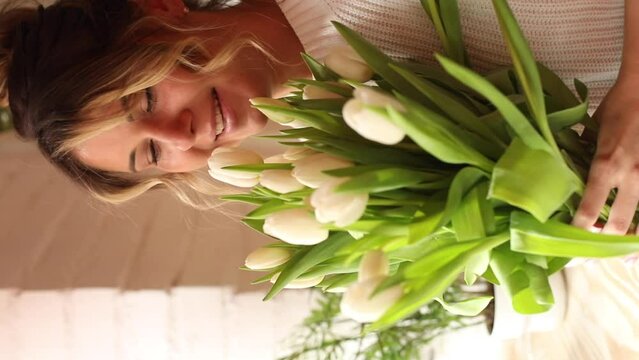 Happy charming young woman enjoys bouquet of white tulips, hugs it to her and sniffs fragrance. Gifts and surprises.
