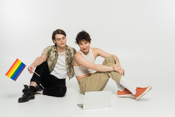 full length of positive bigender couple with small lgbt flag sitting and looking at laptop on grey...