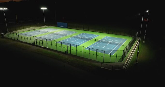 Evening aerial video of outdoor blue tennis courts with pickleball lines with lights turned on.