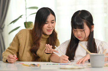 Happy asian mother and little daughter coloring a picture, doing a hobby or education project together at home