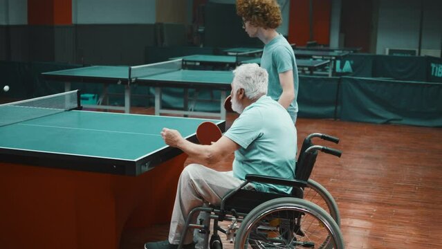 Old man with a disability in a wheelchair serves a ping pong ball, wins a point and rejoices while playing table tennis with his grandson