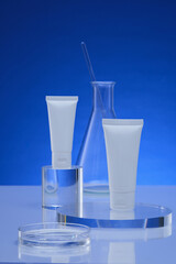 Two white tube without label placed on round and cylinder transparent pedestal, decorated with erlenmeyer flask and petri dish. Empty label for branding mockup