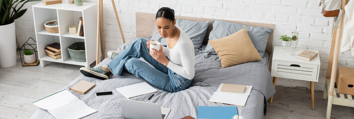 African american student holding tea near devices and notebooks on bed in bedroom, banner.