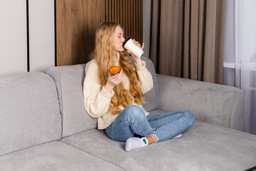 Young beautiful long haired girl in a sweater and jeans sits on a couch with a muffin and a cup of coffee. White mockup paper coffee cup.