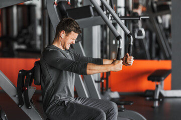 Young strong muscular fit man back muscles workout training with weights on the machine in the gym. The athlete listens to music in earphones.