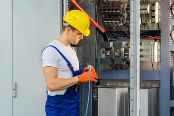 A young electrician in a yellow hard hat cuts the blue wire with pliers. An employee maintains a...