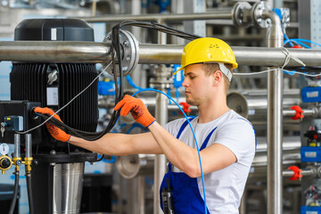 A young worker in a yellow helmet and orange gloves checks the connection of electrical cables at...