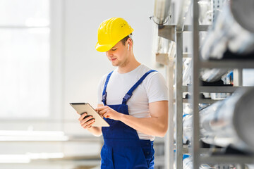 A young smiling industrial worker in yellow hard hat indoors in factory reads technical information from a tablet PC. Control of technical parameters of production.
