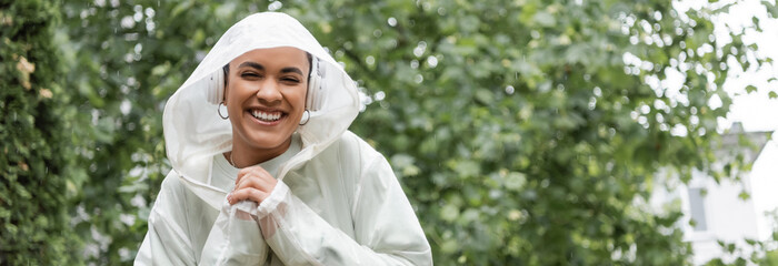 smiling african american woman in waterproof raincoat and wireless headphones standing under rain, banner.