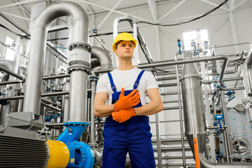 A young worker in a chemical factory wears orange gloves. Metal structures and stainless steel pipes on the background.