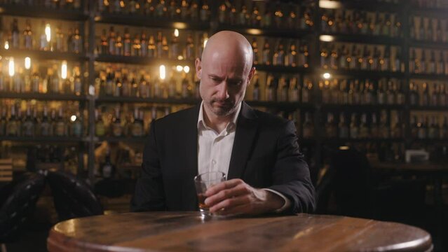 A sad, bald man dressed in a jacket and shirt is sitting in a pub with a glass of whiskey. He looks at the drink with a mournful expression, thinking that things haven't gone right in his life.