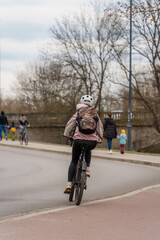 Obraz premium A girl rides a bike in a jacket with a backpack behind her back in a white helmet along the city street on a cloudy day in early spring, eco sustainable urban transport, street photography