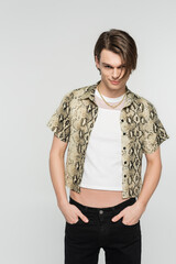 young and trendy bigender model in animal print blouse posing with hands in pockets of black pants isolated on grey.