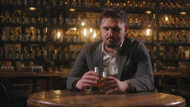 A sad man dressed in a jacket and shirt is sitting in a pub with a glass of whiskey. A disappointed person with a sad expression is reflecting on their life.
