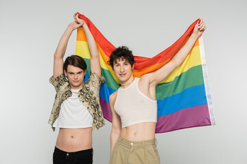 positive nonbinary couple in stylish clothes holding rainbow flag and looking at camera isolated on grey.