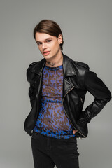 young nonbinary person in animal print top and black leather jacket standing with hands on hips isolated on grey.