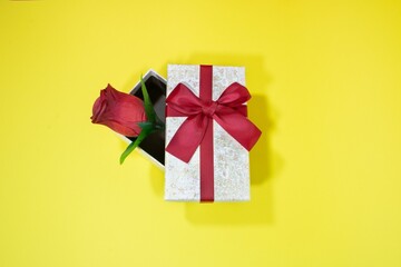 Top view of a small jewellery gift box and an artificial red rose on a yellow surface