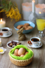 Easter eggs in the basket, bowl of cookies, chocolate pralines, Easter bunny figurine, cups of tea, glasses of juice, flowers and lit candles on the table. Selective focus.