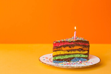 Rainbow birthday cake decorated with a festive candle and confetti. Confectionery. Multicolored dessert in the color of the rainbow on a yellow-orange background.