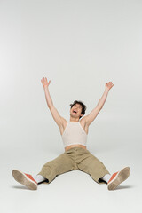 Fototapeta premium excited nonbinary person in beige pants laughing while sitting with raised hands on grey background.